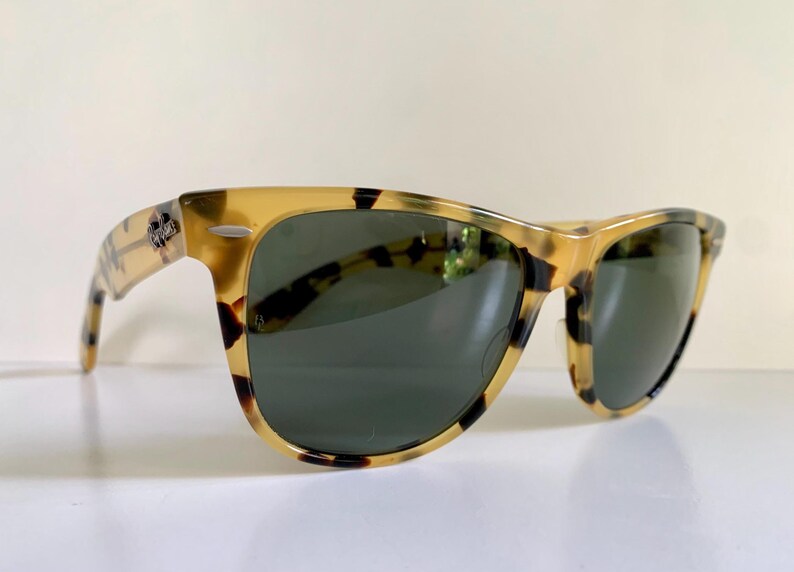 May include: Tortoiseshell sunglasses with dark green lenses. The frames have a classic design with a speckled pattern in shades of brown and yellow. The sunglasses are set against a white background.
