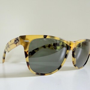 May include: Sunglasses with a classic Wayfarer design. The frames have a tortoiseshell pattern in shades of yellow and brown. The lenses are dark gray, and the Ray-Ban logo is visible on the temple.