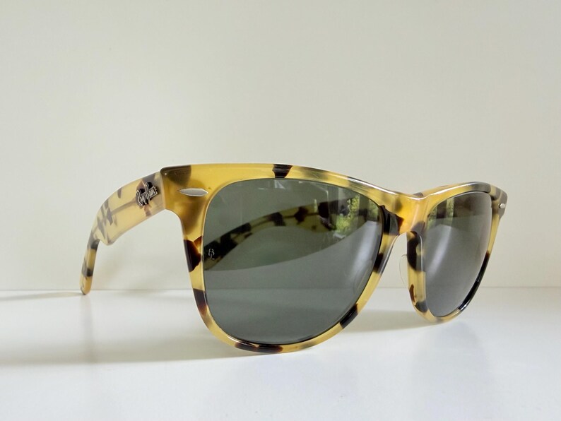 May include: Sunglasses with a classic Wayfarer design. The frames have a tortoiseshell pattern in shades of yellow and brown. The lenses are dark gray, and the Ray-Ban logo is visible on the temple.
