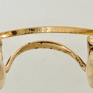 K&ouml;nnte beinhalten: Nahaufnahme einer goldfarbenen Brille mit dem Markennamen "RAY-BAN" auf dem oberen B&uuml;gel eingraviert. Die Brille hat ein klassisches Design mit einem geschwungenen Steg und runden Gl&auml;sern, was auf einen Vintage- oder Retro-Stil hindeutet.