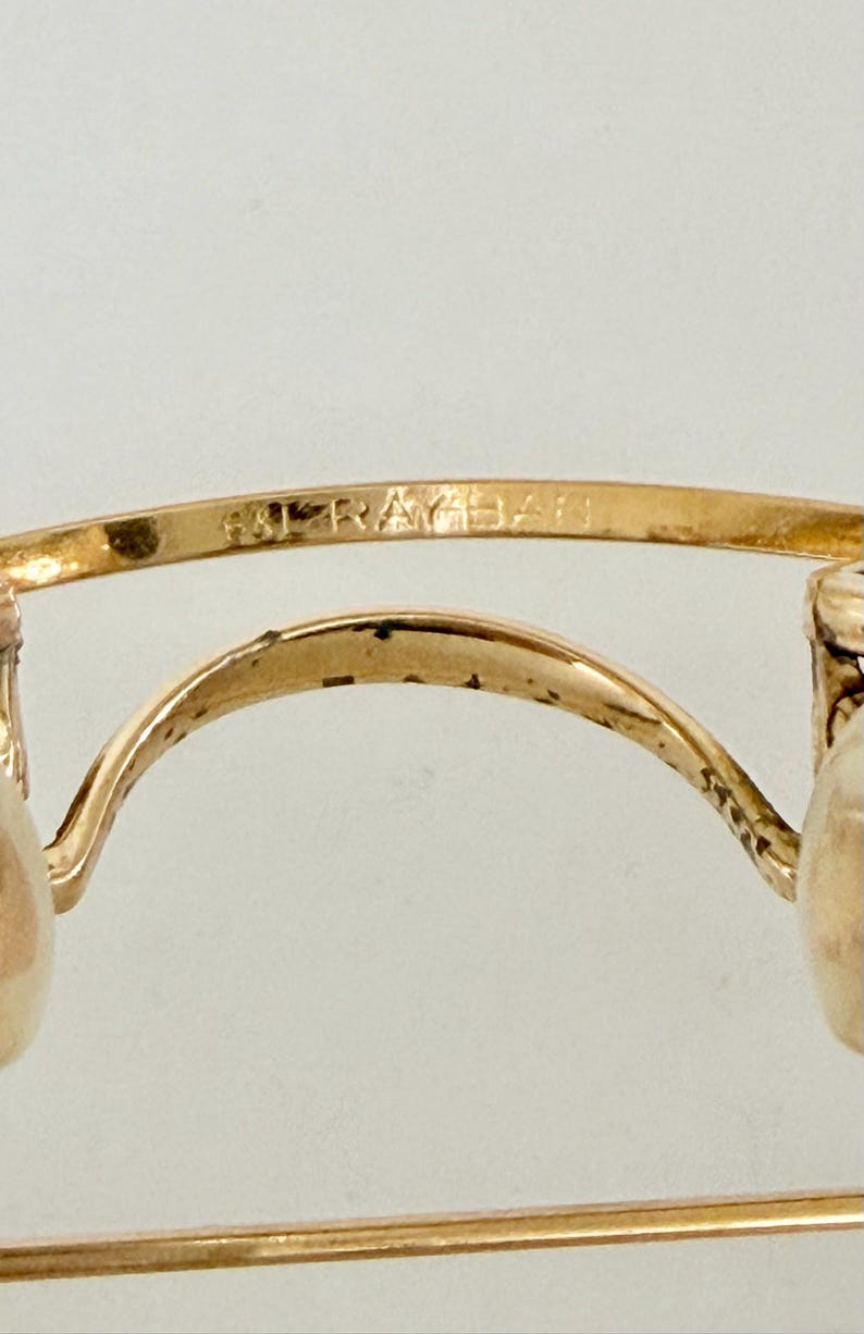 K&ouml;nnte beinhalten: Nahaufnahme einer goldfarbenen Brille mit dem Markennamen "RAY-BAN" auf dem oberen B&uuml;gel eingraviert. Die Brille hat ein klassisches Design mit einem geschwungenen Steg und runden Gl&auml;sern, was auf einen Vintage- oder Retro-Stil hindeutet.