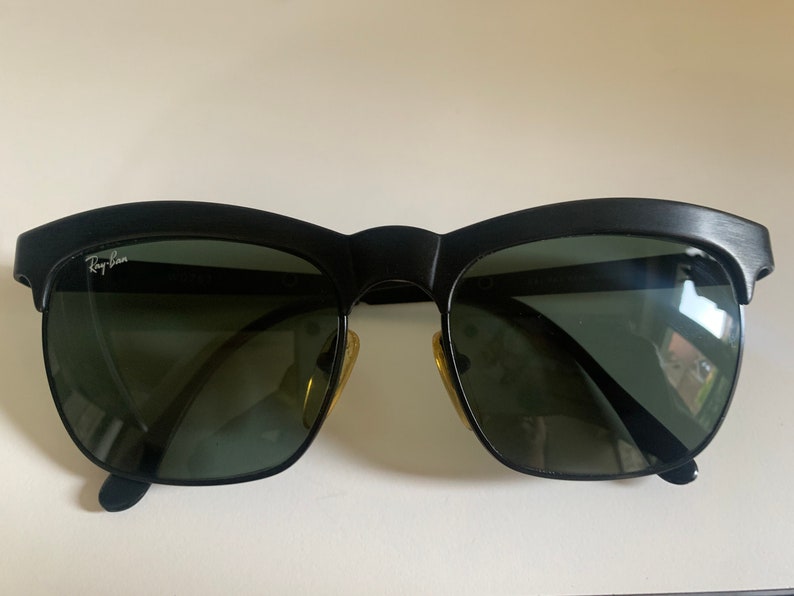 May include: Black sunglasses with a distinctive browline design. The sunglasses feature dark lenses and a matte black frame. The Ray-Ban logo is visible on the upper left lens. The sunglasses have a classic, retro style.