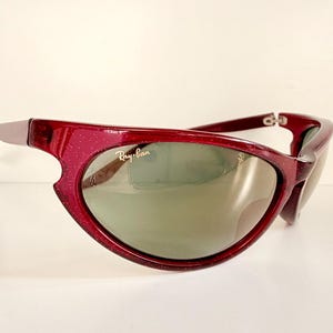 May include: A pair of Ray-Ban sunglasses with a deep red frame and green-tinted lenses. The frame has a unique, curved design with a speckled texture. The Ray-Ban logo is visible on the lens.