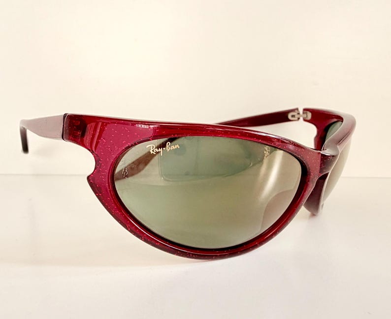 May include: A pair of Ray-Ban sunglasses with a deep red frame and green-tinted lenses. The frame has a unique, curved design with a speckled texture. The Ray-Ban logo is visible on the lens.