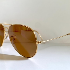 May include: Gold-coloured aviator sunglasses with brown lenses. The sunglasses feature a double bridge and a classic teardrop shape. The temples are thin and extend to the ear pieces. The lenses are a warm brown colour, and the frame is a shiny gold.