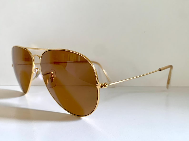 May include: Gold-coloured aviator sunglasses with brown lenses. The sunglasses feature a double bridge and a classic teardrop shape. The temples are thin and extend to the ear pieces. The lenses are a warm brown colour, and the frame is a shiny gold.