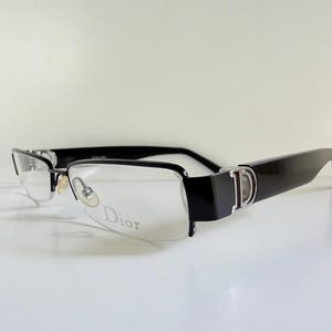 Könnte beinhalten: Schwarze rechteckige Brille mit klaren Gläsern. Die Fassungen haben ein silberfarbenes 'D'-Logo an den Bügeln und das Wort 'Dior' ist auf dem Glas aufgedruckt. Die Brille hat ein schlankes, modernes Design.