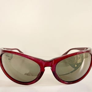 May include: Burgundy-colored sunglasses with a thick, rounded frame and dark lenses. The sunglasses have a slight sparkle and a curved design. The lenses are a dark gray color, and the arms are a matching burgundy.