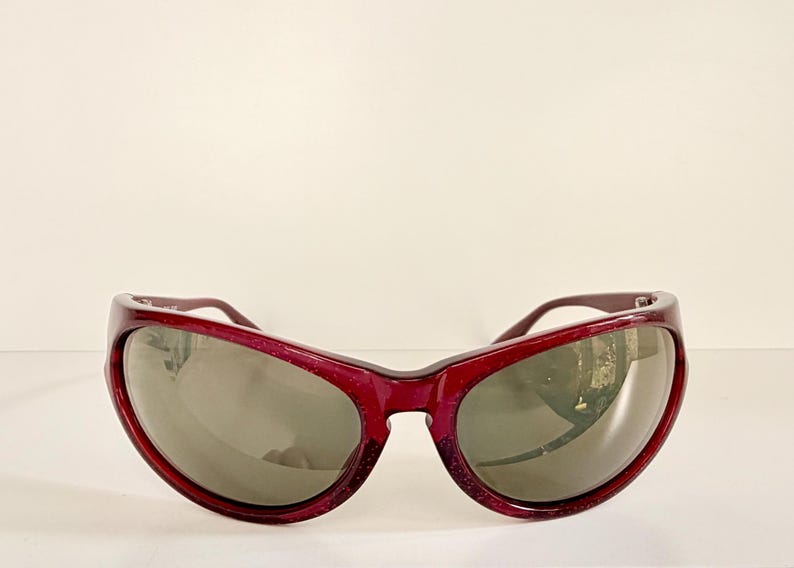 May include: Burgundy-colored sunglasses with a thick, rounded frame and dark lenses. The sunglasses have a slight sparkle and a curved design. The lenses are a dark gray color, and the arms are a matching burgundy.