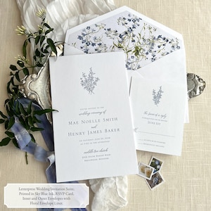 May include: A white wedding invitation suite with blue floral accents. The invitation features the names "Mae Noelle Smith" and "Henry James Baker" with the wedding date and location. The RSVP card includes the text "the favor of a reply is requested on or before June 1st".