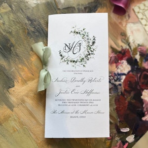 May include: A wedding program with a floral wreath design and the initials "NJ". The program is tied with a light green ribbon. The text includes the names Nadine Dorothy Roberts and Justin Eric Hoffman, and the date.