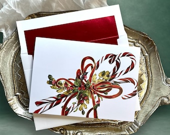 May include: A white greeting card featuring a watercolor illustration of two candy canes crossed behind a festive arrangement of red ribbons, green holly leaves, and small red berries. The card is tucked into a white envelope with a red inner lining.