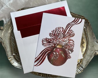 May include: A white greeting card featuring a watercolor illustration of a red Christmas ornament with gold accents and a large, flowing red and white striped ribbon bow. The card is paired with a white envelope that has a deep red inner lining.