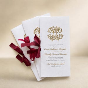 May include: Three white wedding programs with gold foil monograms and text. The text reads: The Celebration of Marriage Uniting Carrie Catherine Wampler and Timothy Connor Alexander. Saturday, the first of October, two thousand twenty-two. Deep red ribbons.