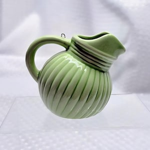 May include: A small, light green ceramic pitcher with a rounded body and a handle. The pitcher has a ribbed design on the body and a spout. The handle is curved, and there is a small loop at the top for hanging. The pitcher is likely a decorative item.