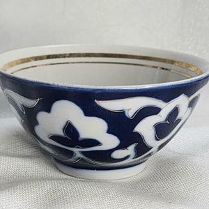 May include: A ceramic bowl with a white interior and a navy blue exterior. The bowl features a gold rim and a repeating white floral pattern outlined in gold. The bowl is likely used for serving food or as a decorative piece.