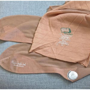 Peut inclure: Bas de nylon couleur beige avec l'inscription "ISIS NYLONS" et "Right Angle Foot". Les bas sont pliés, révélant les parties du pied et de la jambe. On distingue également "51 gauge" et la taille "D 10".
