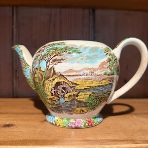Puede incluir: Una tetera de cerámica vintage con una escena de paisaje que representa un molino de agua, árboles y un río. La tetera tiene un asa y un pico curvos, y un estampado floral en la base. Los colores incluyen verde, marrón, azul y amarillo.