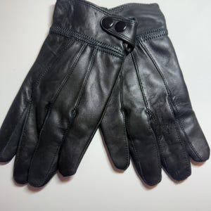 May include: A pair of black leather gloves featuring a smooth finish, stitched detailing along the fingers, and a wrist strap secured with two black snap buttons. The gloves are positioned side by side, showcasing the palm and finger construction.