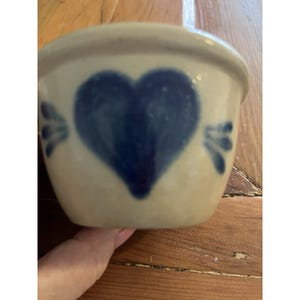 Puede incluir: Un pequeño cuenco de cerámica blanquecina con un gran corazón azul pintado en el frente. Dos diseños florales azules a cada lado del corazón. El cuenco tiene forma redondeada y un borde ligeramente ensanchado.