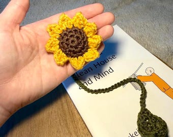 May include: A handmade crochet sunflower with yellow petals and a brown center. The sunflower is held in a hand, next to a book with the title "Lean House and Mind" and a green crochet bookmark.