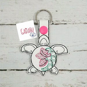 May include: A white and black embroidered turtle keychain with a pink button and silver keyring. The turtle design features a geometric pattern in red and green. A small white tag with the text "GADAHU" is attached.