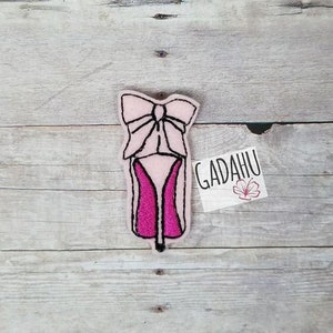 May include: A pink and fuchsia high heel shoe-shaped felt applique with a bow at the top. The applique is outlined in black stitching. A small white tag with the text "GADAHU" is on the right.