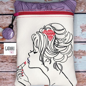 Könnte beinhalten: Eine weiße Kosmetiktasche mit einer schwarzen Umrisszeichnung einer Frau, die Lippenstift aufträgt. Die Tasche hat eine lila, floral gemusterte Oberseite, einen roten Reißverschluss und einen lila Anhänger. Der Markenname "GADAHU" ist auf einem weißen Etikett aufgedruckt.