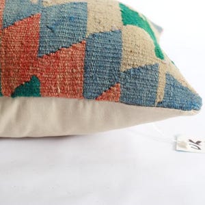 K&ouml;nnte beinhalten: Ein rechteckiges Zierkissen mit einer gewebten Vorderseite im Kelim-Stil mit geometrischem Muster in Terrakotta, Blau, Gr&uuml;n und Creme. Die R&uuml;ckseite des Kissens besteht aus einfarbigem, cremefarbenem Stoff. Ein kleines wei&szlig;es Etikett ist befestigt.