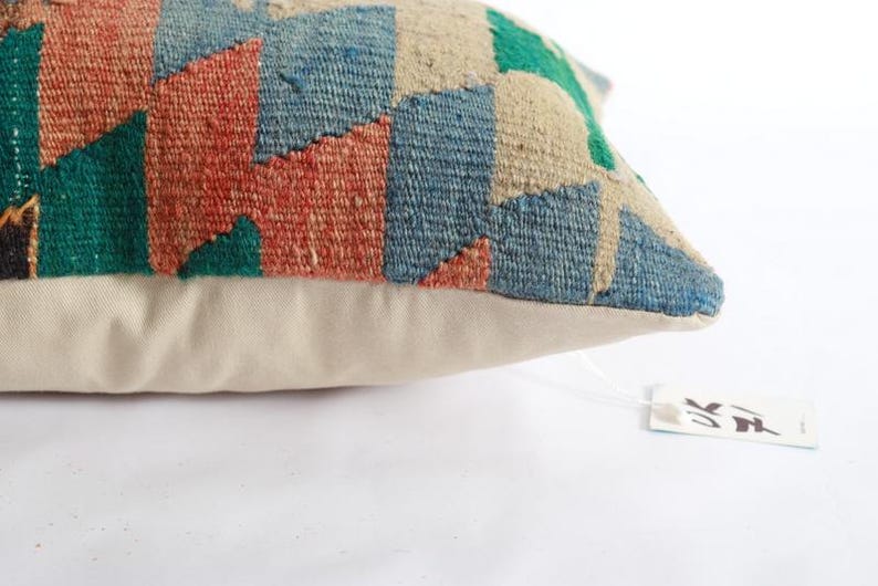 K&ouml;nnte beinhalten: Ein rechteckiges Zierkissen mit einer gewebten Vorderseite im Kelim-Stil mit geometrischem Muster in Terrakotta, Blau, Gr&uuml;n und Creme. Die R&uuml;ckseite des Kissens besteht aus einfarbigem, cremefarbenem Stoff. Ein kleines wei&szlig;es Etikett ist befestigt.