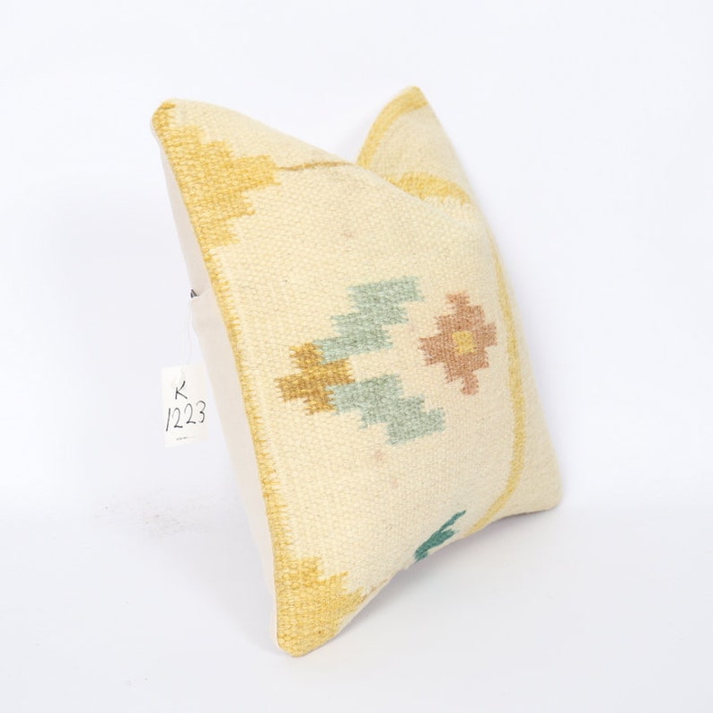 May include: Cream-coloured decorative cushion with geometric patterns in gold, light blue, and peach. The cushion has a textured surface and a solid cream-coloured back. The tag reads "K 1223".