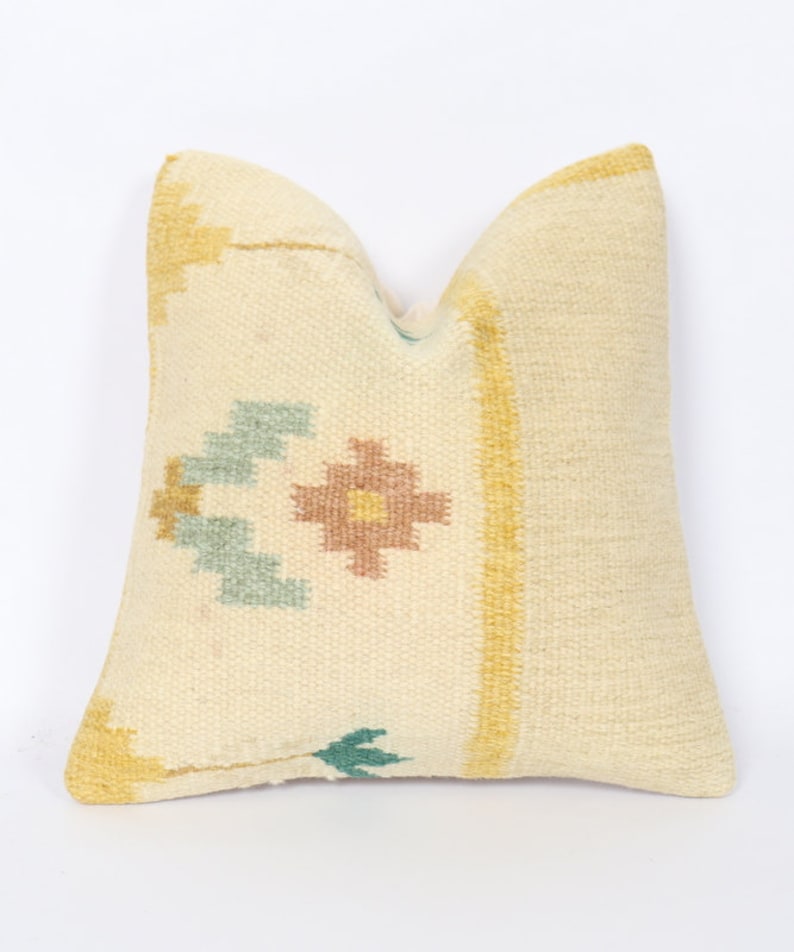 May include: Cream-coloured decorative cushion with a woven texture. The cushion features a central design with a floral motif in shades of brown, blue, and yellow. Yellow stripes border the cushion's edges, adding colour to the neutral base.