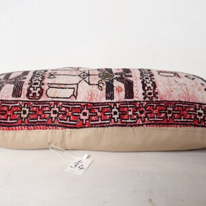 Puede incluir: Una almohada rectangular con fondo blanco y un patr&oacute;n geom&eacute;trico rojo, negro y blanco. La almohada tiene un respaldo de tela beige y una etiqueta blanca con el n&uacute;mero "34" en ella.