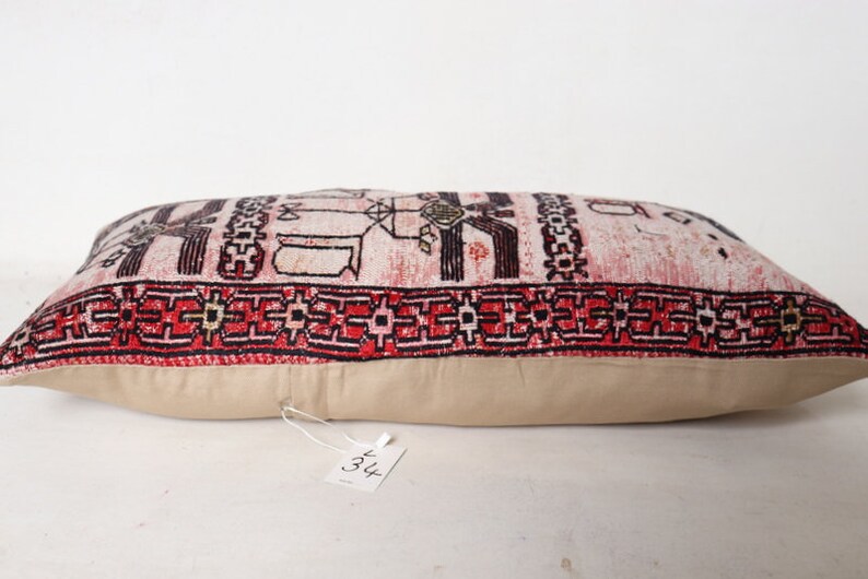Puede incluir: Una almohada rectangular con fondo blanco y un patr&oacute;n geom&eacute;trico rojo, negro y blanco. La almohada tiene un respaldo de tela beige y una etiqueta blanca con el n&uacute;mero "34" en ella.