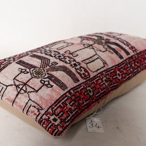 Puede incluir: Una almohada rectangular con una tela estampada en rosa y rojo. La tela presenta un dise&ntilde;o geom&eacute;trico con acentos en negro y blanco. La almohada est&aacute; hecha de un material suave y tejido y tiene un respaldo beige.