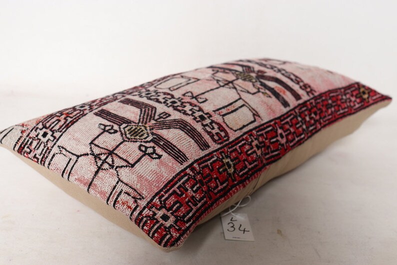 Puede incluir: Una almohada rectangular con una tela estampada en rosa y rojo. La tela presenta un dise&ntilde;o geom&eacute;trico con acentos en negro y blanco. La almohada est&aacute; hecha de un material suave y tejido y tiene un respaldo beige.