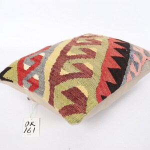 May include: A colorful, rectangular throw pillow with a geometric pattern in shades of red, green, black, and gray. The pillow has a beige backing and a white tag with the text "OK 161" on it.