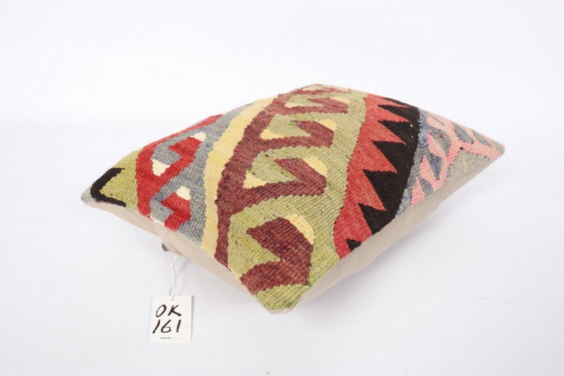 May include: A colorful, rectangular throw pillow with a geometric pattern in shades of red, green, black, and gray. The pillow has a beige backing and a white tag with the text "OK 161" on it.