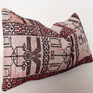 Puede incluir: Una almohada rectangular con fondo blanco y un patr&oacute;n geom&eacute;trico rojo, negro y verde. La almohada est&aacute; hecha de tejido tejido y tiene un borde con flecos.