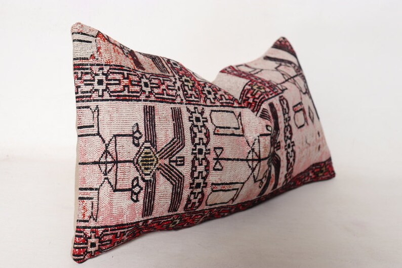 Puede incluir: Una almohada rectangular con fondo blanco y un patr&oacute;n geom&eacute;trico rojo, negro y verde. La almohada est&aacute; hecha de tejido tejido y tiene un borde con flecos.