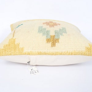 May include: Cream-coloured decorative cushion with a geometric design in yellow, light blue, and pink. The cushion has a soft, textured surface and a white backing. The tag reads "1223".