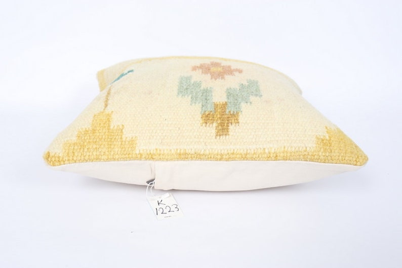 May include: Cream-coloured decorative cushion with a geometric design in yellow, light blue, and pink. The cushion has a soft, textured surface and a white backing. The tag reads "1223".