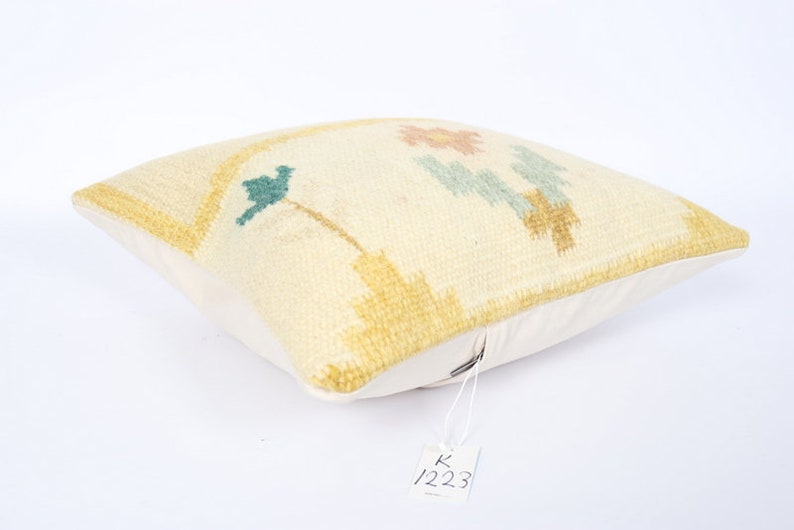 May include: Cream-coloured square cushion with a floral design in shades of yellow, teal, and peach. The cushion has a textured surface and a white backing. A tag with the number 1223 is attached.