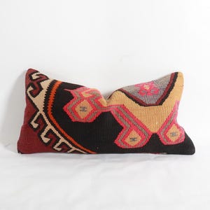 Puede incluir: Cojín decorativo rectangular con un patrón geométrico. El cojín presenta un fondo negro con formas geométricas rojas, beige, naranjas y grises. El cojín está hecho de tela tejida y probablemente se utiliza para la decoración del hogar.