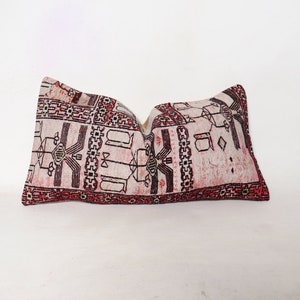 Puede incluir: Una almohada rectangular con fondo blanco y un patr&oacute;n geom&eacute;trico rojo, negro y blanco. La almohada est&aacute; hecha de una tela tejida y tiene un borde con flecos.