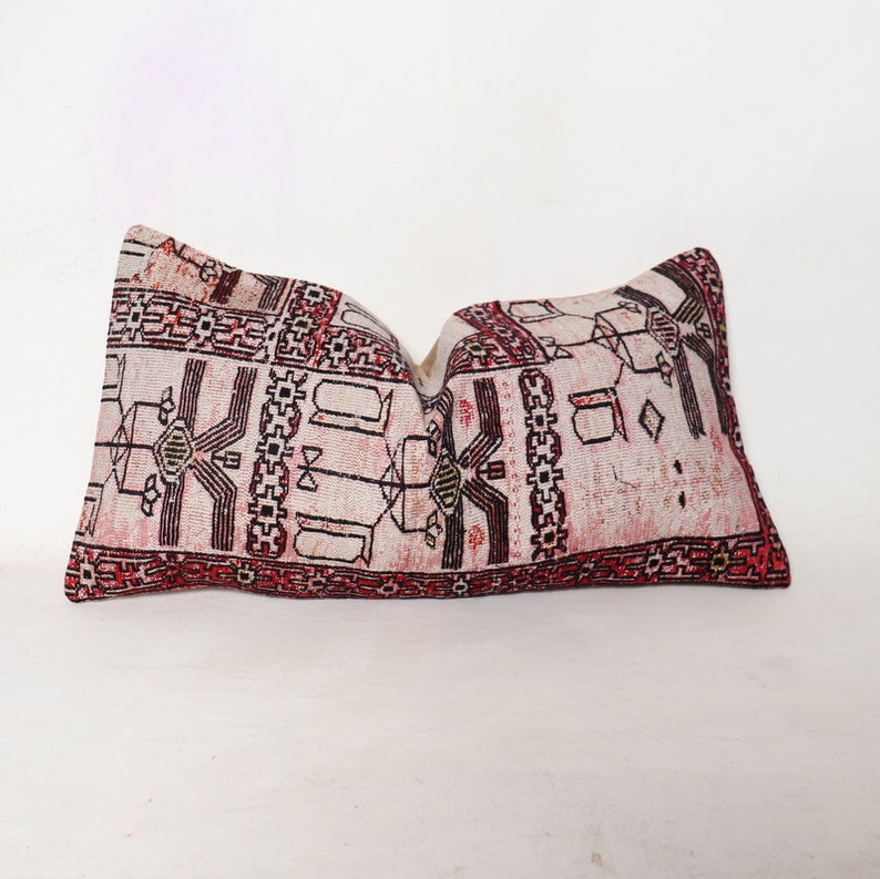 Puede incluir: Una almohada rectangular con fondo blanco y un patr&oacute;n geom&eacute;trico rojo, negro y blanco. La almohada est&aacute; hecha de una tela tejida y tiene un borde con flecos.