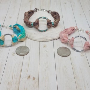 K&ouml;nnte beinhalten: Drei Seidenarmb&auml;nder mit silbernen Kreisanh&auml;ngern. Die Armb&auml;nder sind in Braun-, Rosa- und Blaut&ouml;nen gehalten.