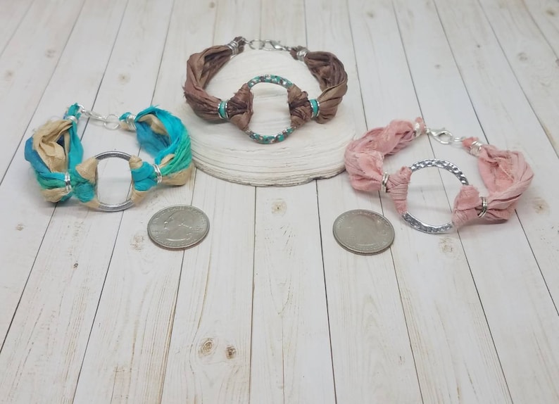 K&ouml;nnte beinhalten: Drei Seidenarmb&auml;nder mit silbernen Kreisanh&auml;ngern. Die Armb&auml;nder sind in Braun-, Rosa- und Blaut&ouml;nen gehalten.