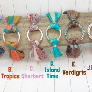K&ouml;nnte beinhalten: Sechs silberne Reifenarmb&auml;nder mit verschiedenen farbigen Seidenband-Quasten. Die Quasten sind in den Farben Silber, Orange, Rosa, T&uuml;rkis, Braun und Rosa. Die Armb&auml;nder sind mit A. Silver Moon, B. Tropics, C. Sherbert, D. Island Time, E. Verdigris und F. Blushing beschriftet.