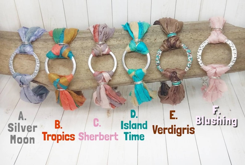K&ouml;nnte beinhalten: Sechs silberne Reifenarmb&auml;nder mit verschiedenen farbigen Seidenband-Quasten. Die Quasten sind in den Farben Silber, Orange, Rosa, T&uuml;rkis, Braun und Rosa. Die Armb&auml;nder sind mit A. Silver Moon, B. Tropics, C. Sherbert, D. Island Time, E. Verdigris und F. Blushing beschriftet.